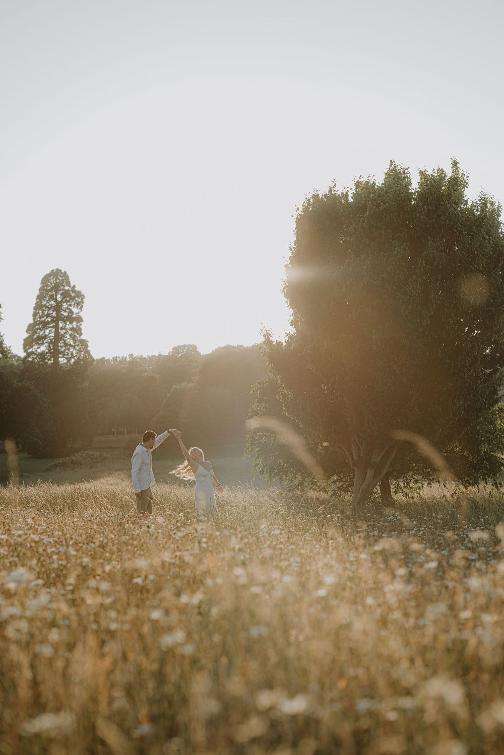 Couple dances in golden meadow during sunset with trees creating dramatic backdrop.