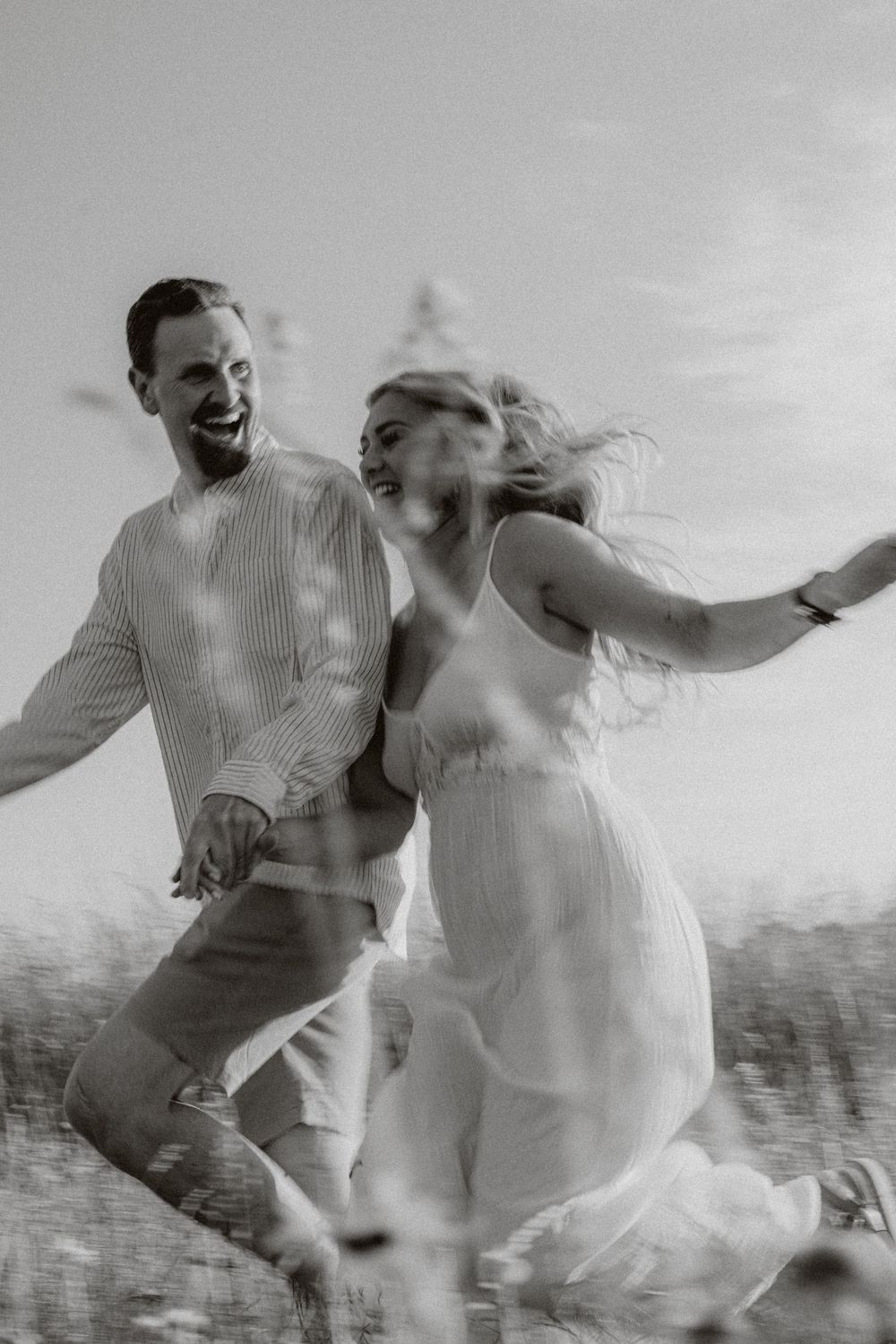 Black and white photo captures joyful couple dancing and laughing in meadow.