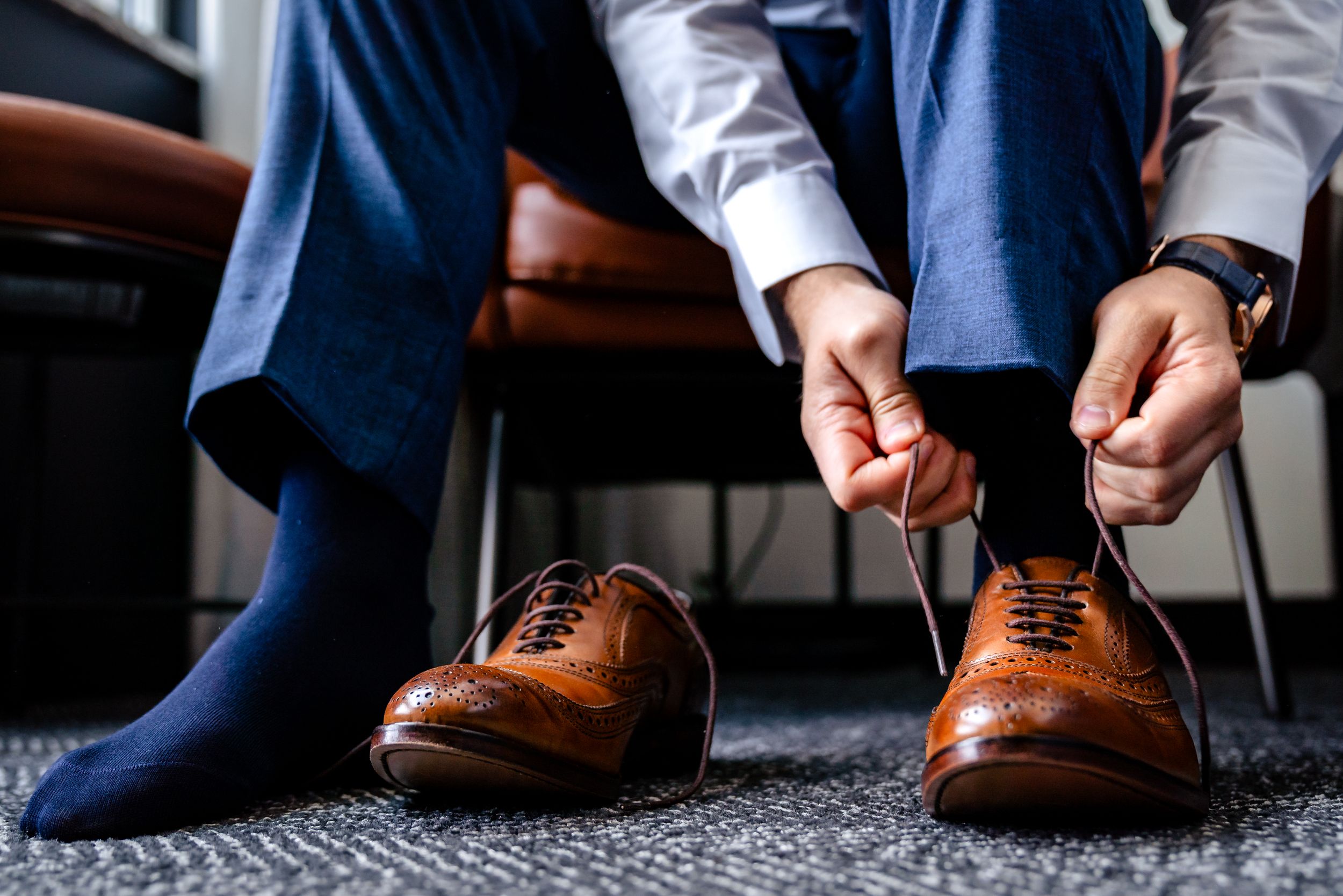 Close-up of hands tying brown leather dress shoes with blue suit pants and socks.