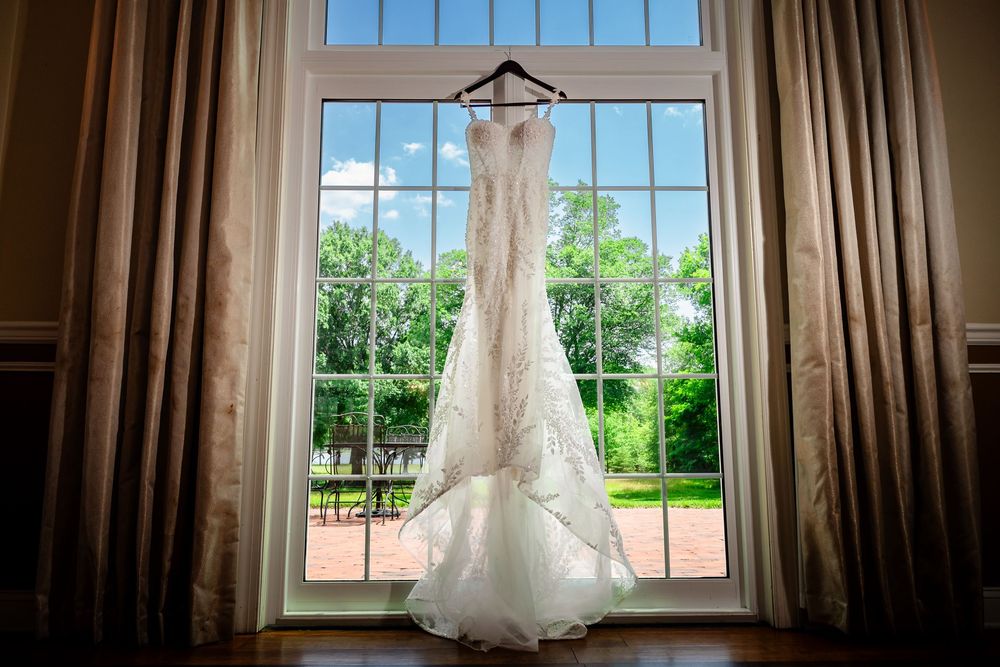 White lace wedding dress hanging in window with natural light streaming through.