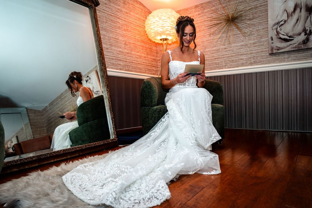 A person in a white wedding dress sits elegantly in front of a vintage mirror with warm lighting.