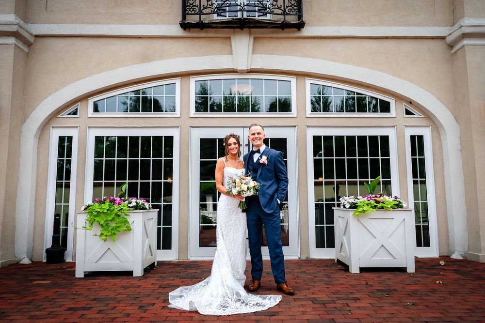 Wedding party poses in front of elegant white arched doorway with brick walkway.