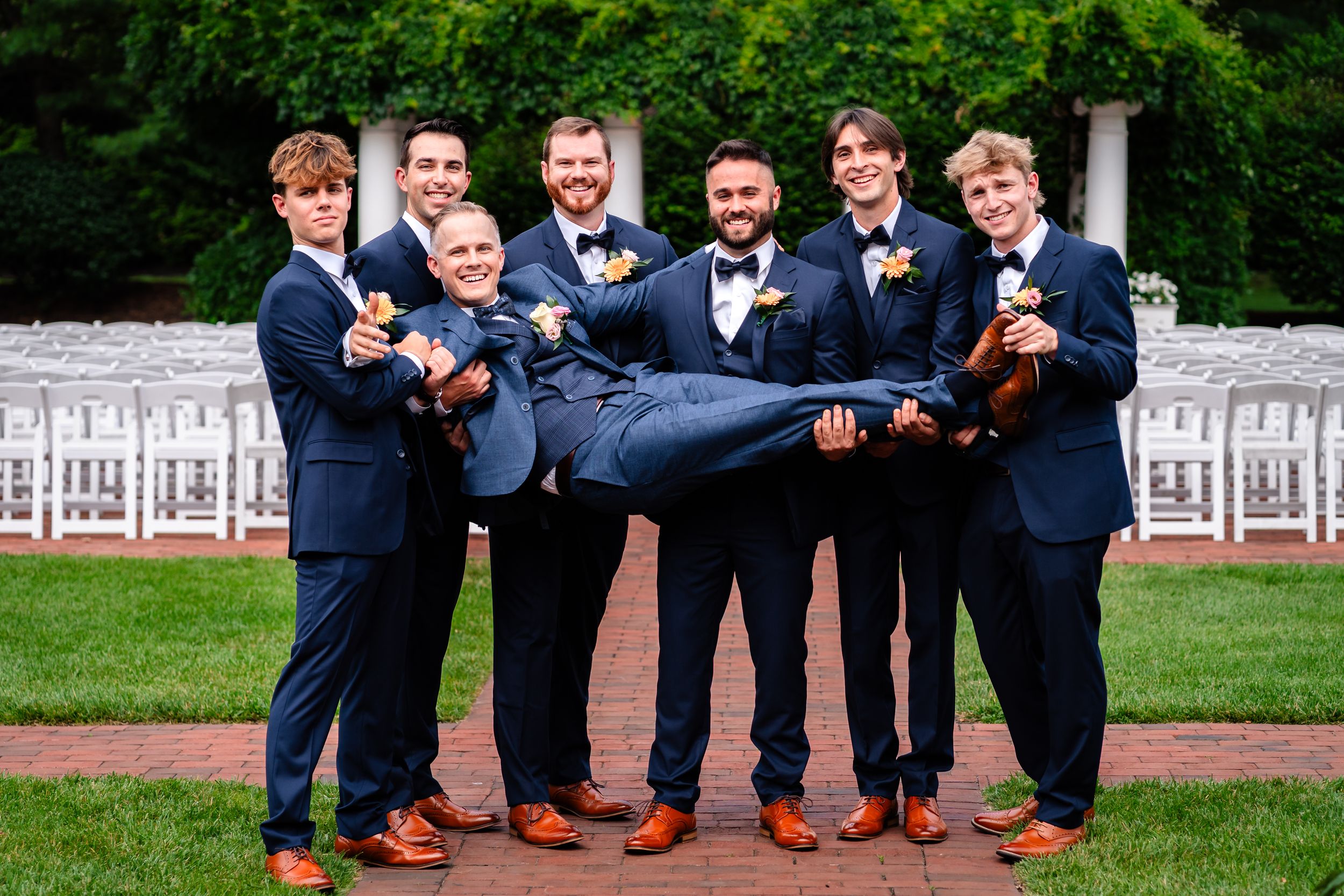 Groomsmen in navy suits pose playfully on brick patio with white chairs in background.
