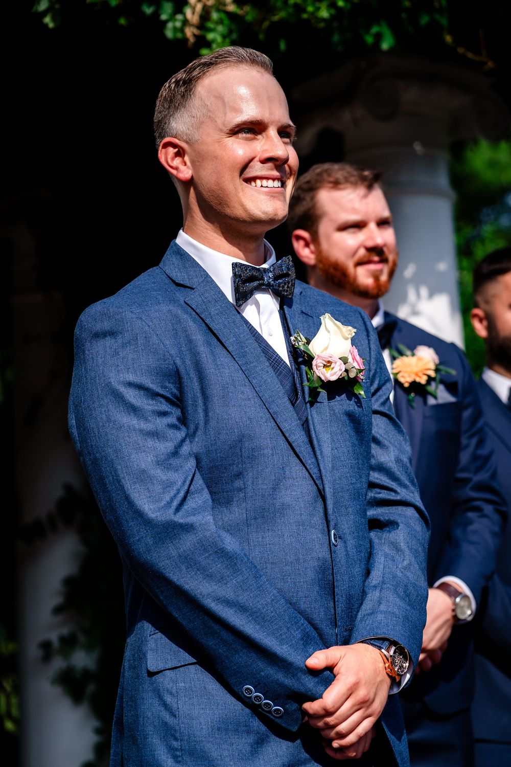 Groomsmen in navy blue suits standing together during outdoor wedding ceremony.
