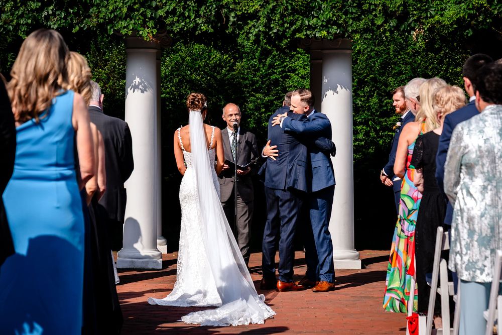 Wedding party and guests gather on a brick patio surrounded by white columns and lush greenery during an outdoor ceremony.