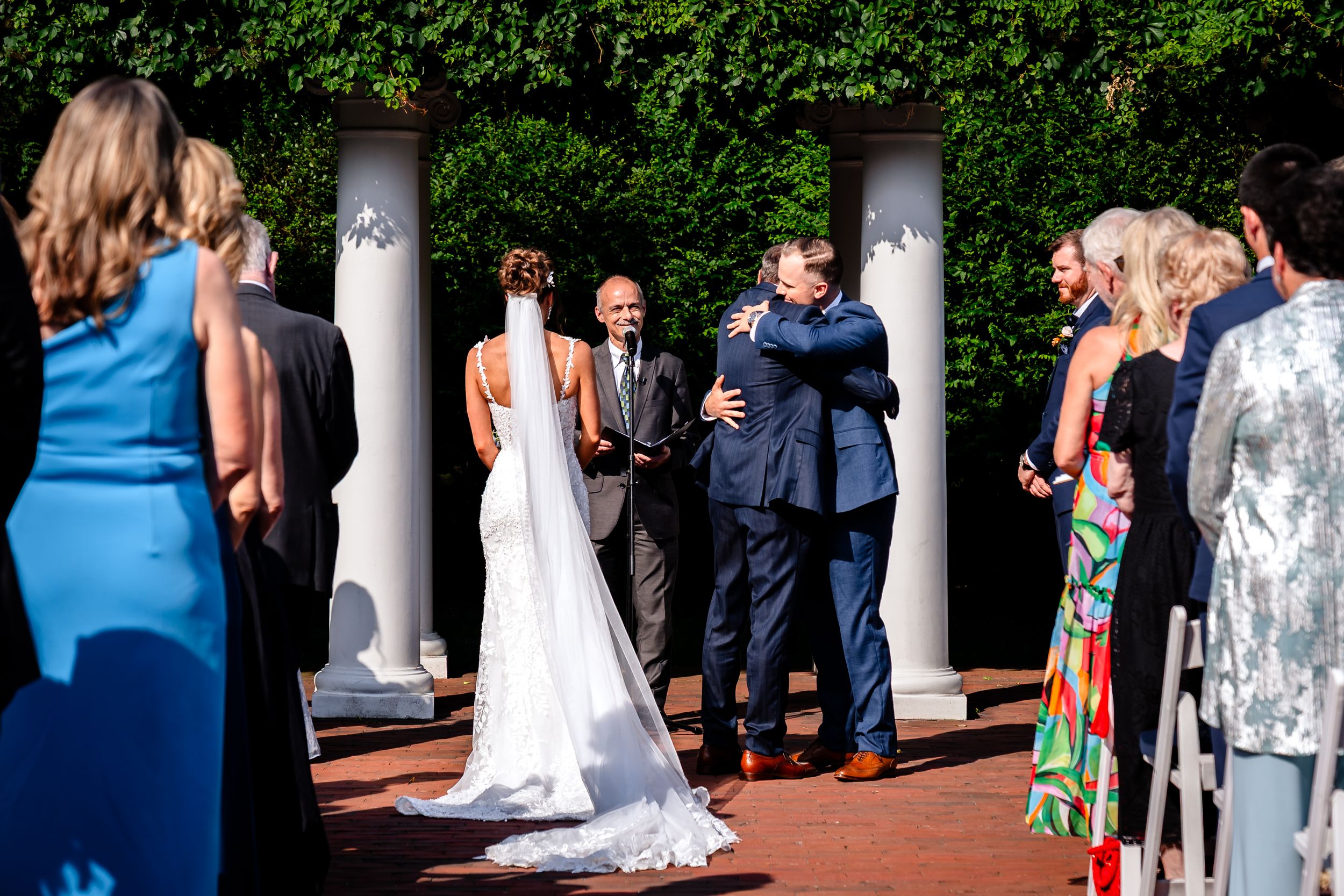 Wedding party and guests gather on a brick patio surrounded by white columns and lush greenery during an outdoor ceremony.