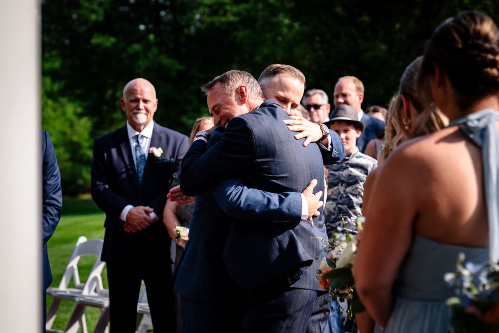 Wedding guests mingle and socialize during an outdoor reception with white columns and greenery in the background.