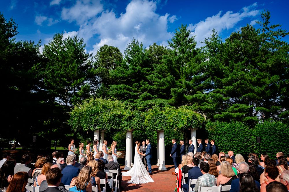 Panoramic view of an outdoor wedding venue with white chairs, columns and pine trees under blue summer skies.
