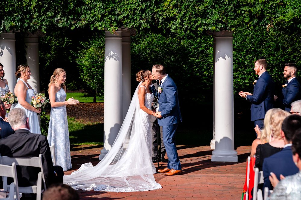 Wedding party poses between elegant white columns along a brick walkway surrounded by lush foliage.