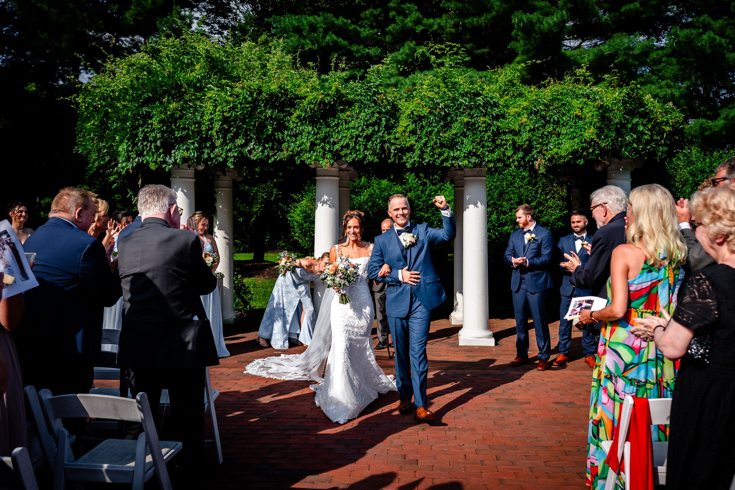 Guests gather on a brick patio during an outdoor wedding reception surrounded by greenery and columns.