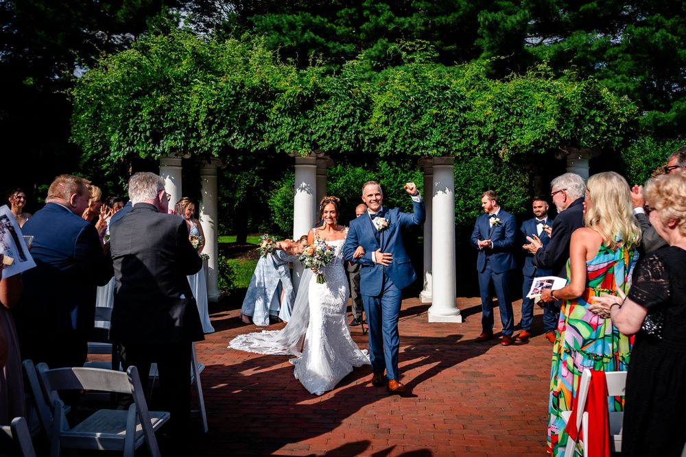 Guests gather on a brick patio during an outdoor wedding reception surrounded by greenery and columns.