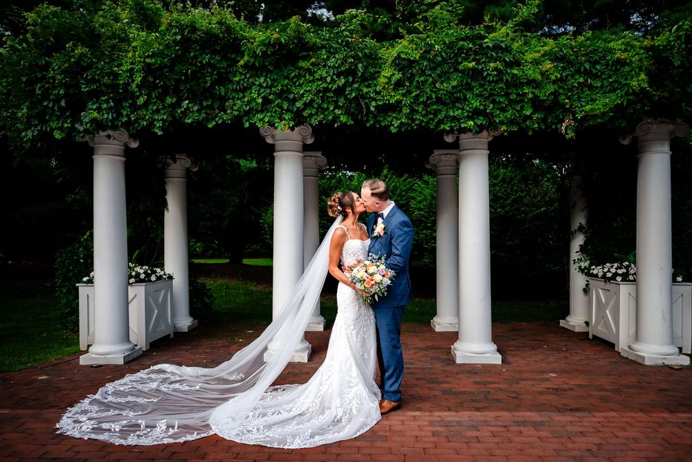 A romantic wedding couple poses between elegant white columns on a brick patio with flowing veil and formal attire.
