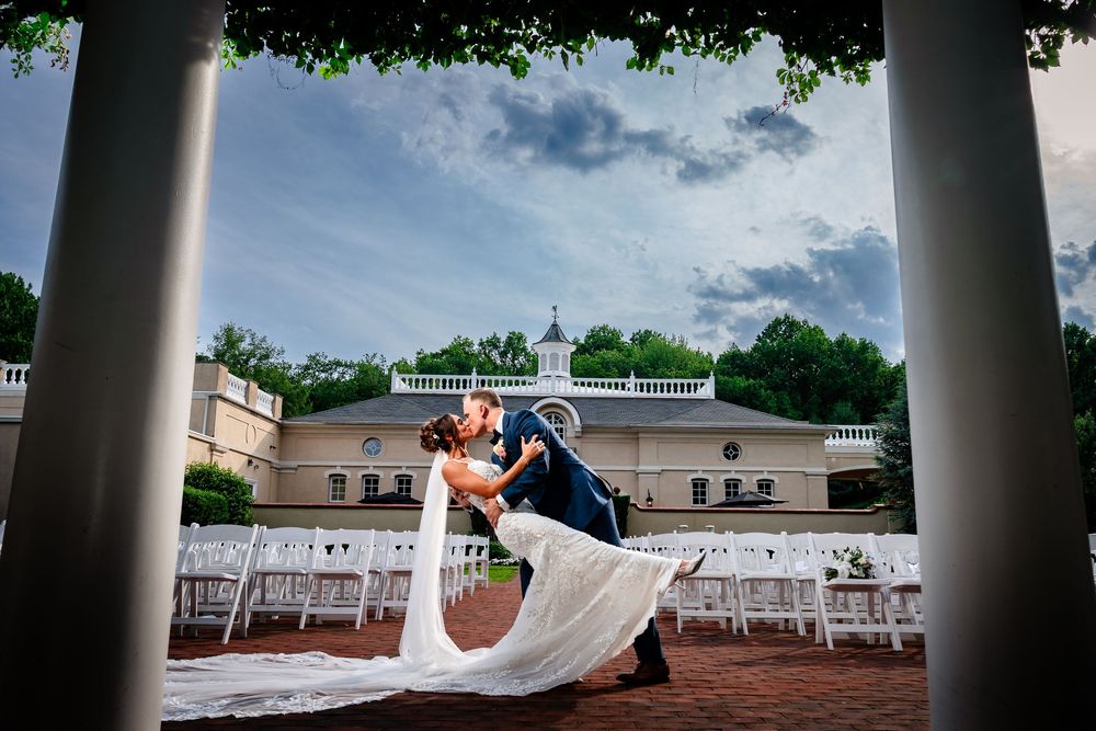 Wedding couple embraces under a colonnade of white pillars against a bright blue summer sky.