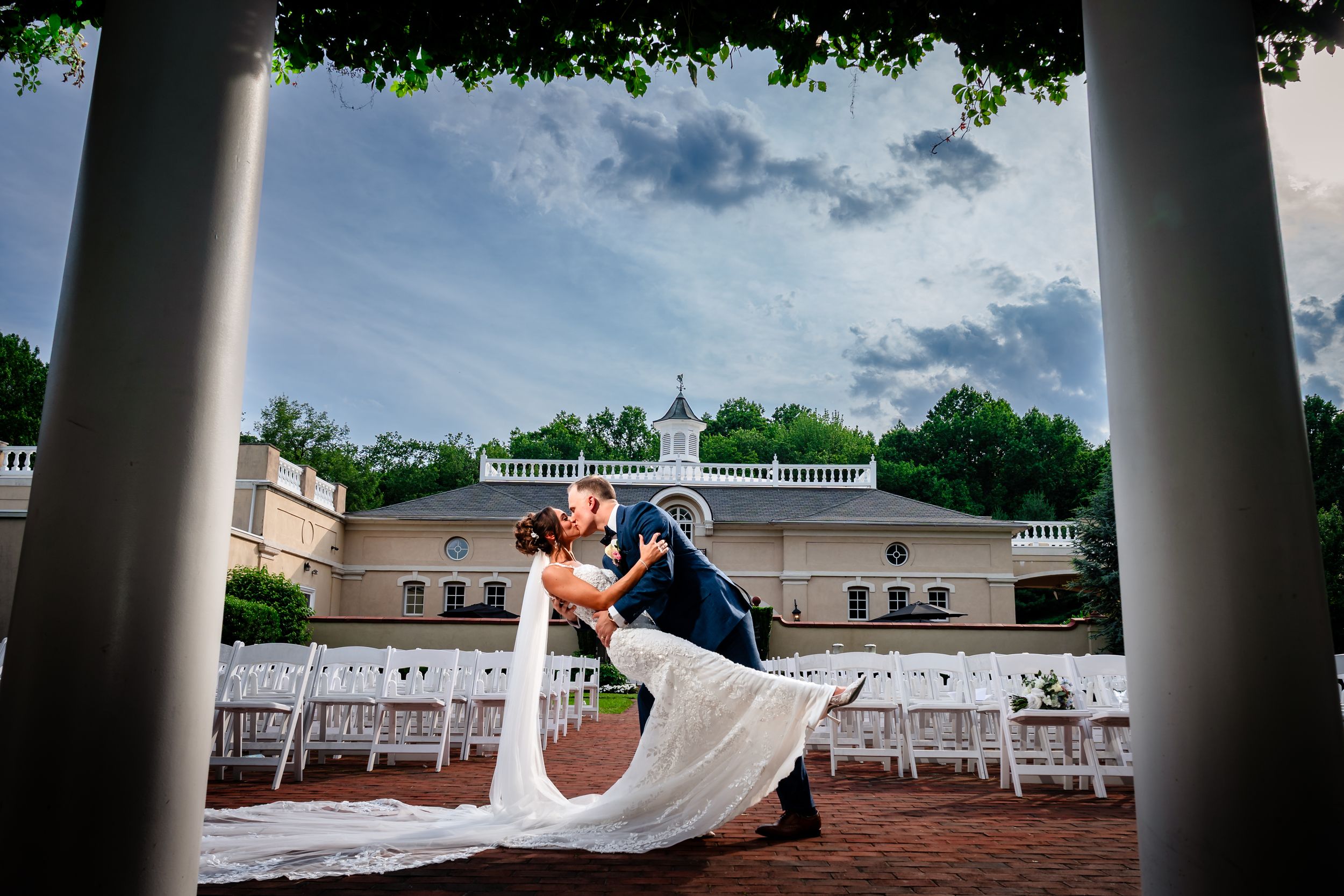 Wedding couple embraces under a colonnade of white pillars against a bright blue summer sky.