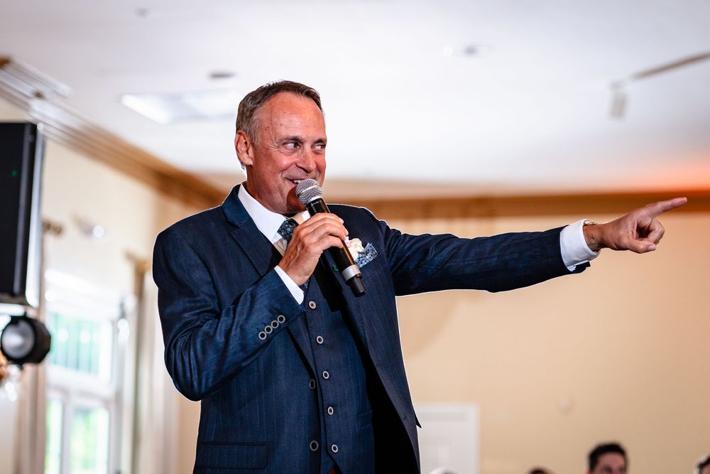 Wedding reception speaker gestures enthusiastically while addressing the crowd at an indoor venue.