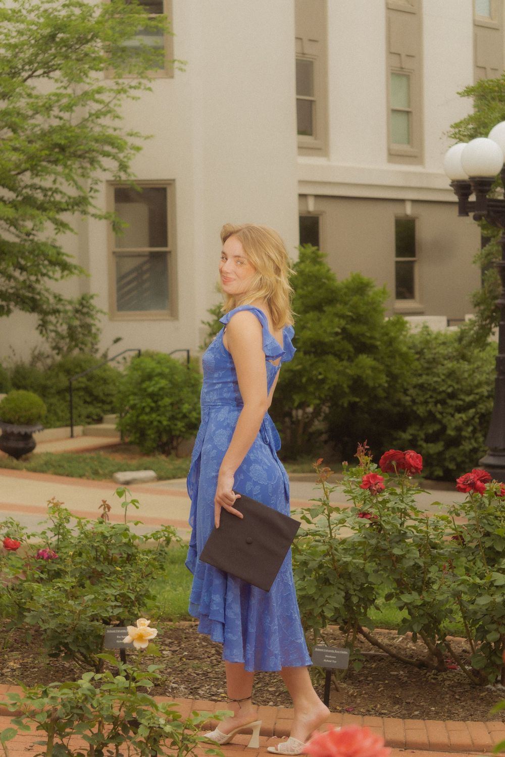 Graduate poses for portraits near blooming rose bushes outside a campus building.