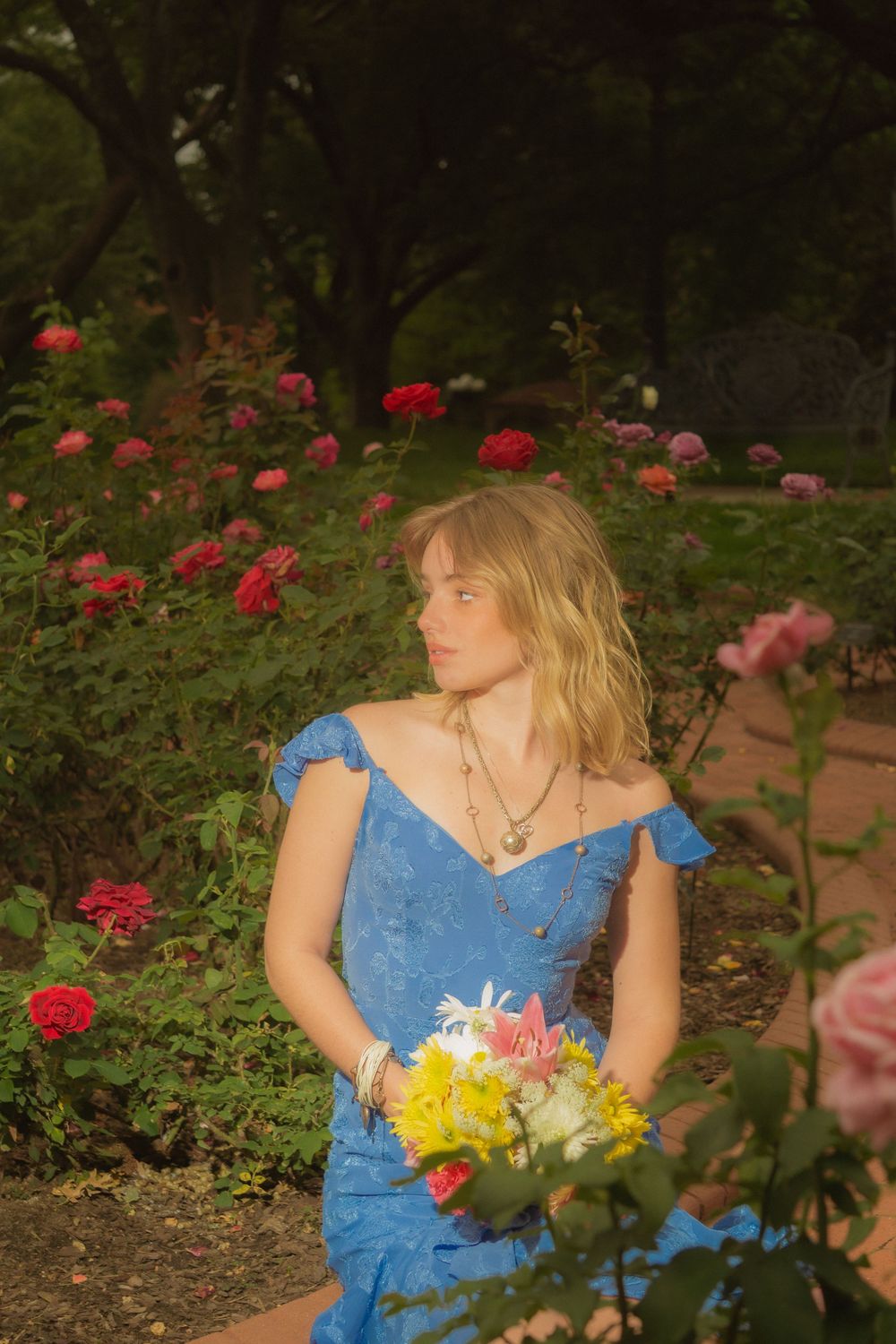 Graduate in blue dress poses with colorful flowers in a rose garden setting.