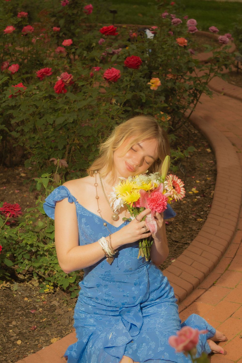 Graduate in blue dress enjoys a moment with fresh flowers in a garden setting.