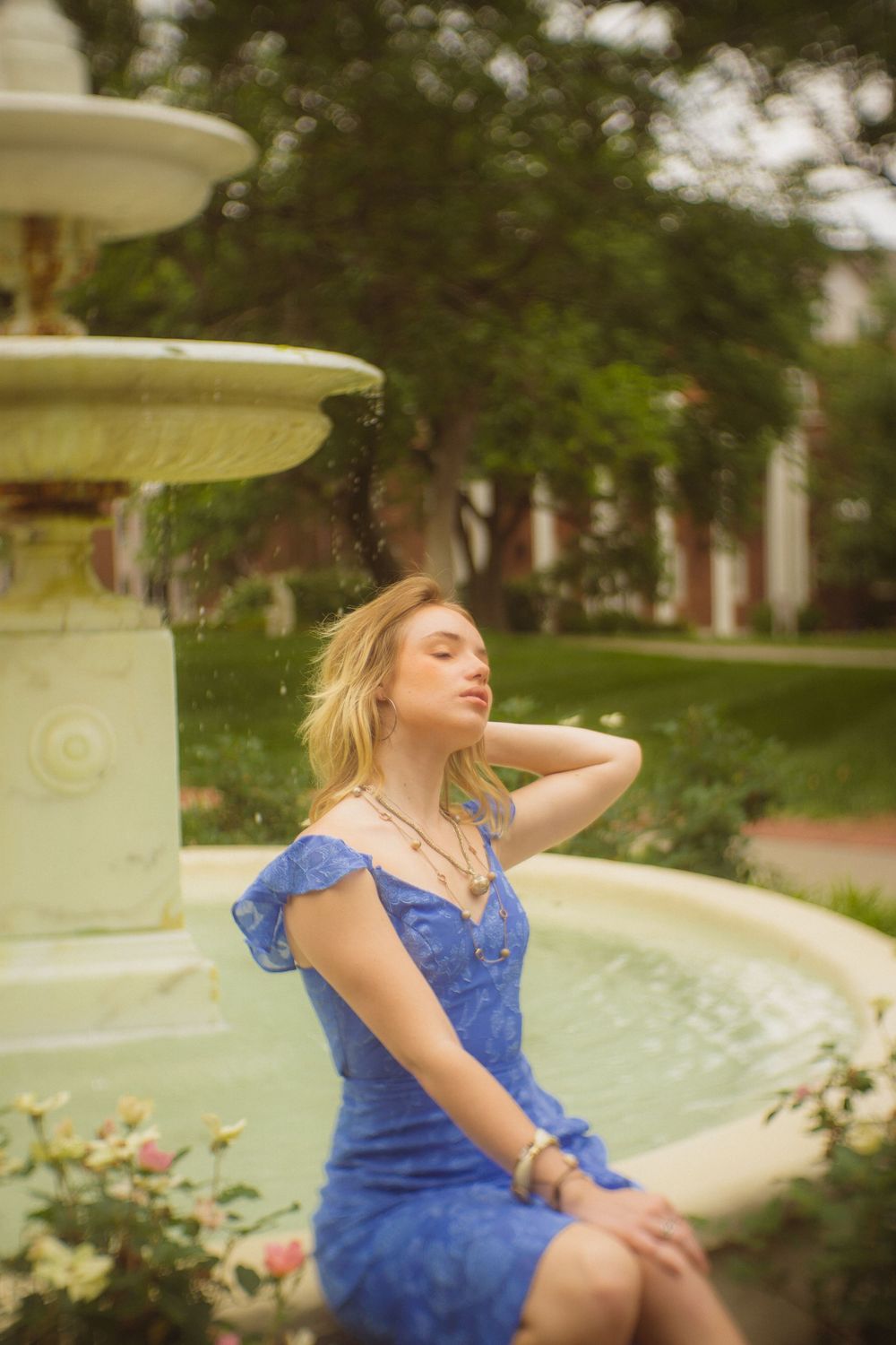 Graduate in blue dress poses artistically by campus fountain during photo session.