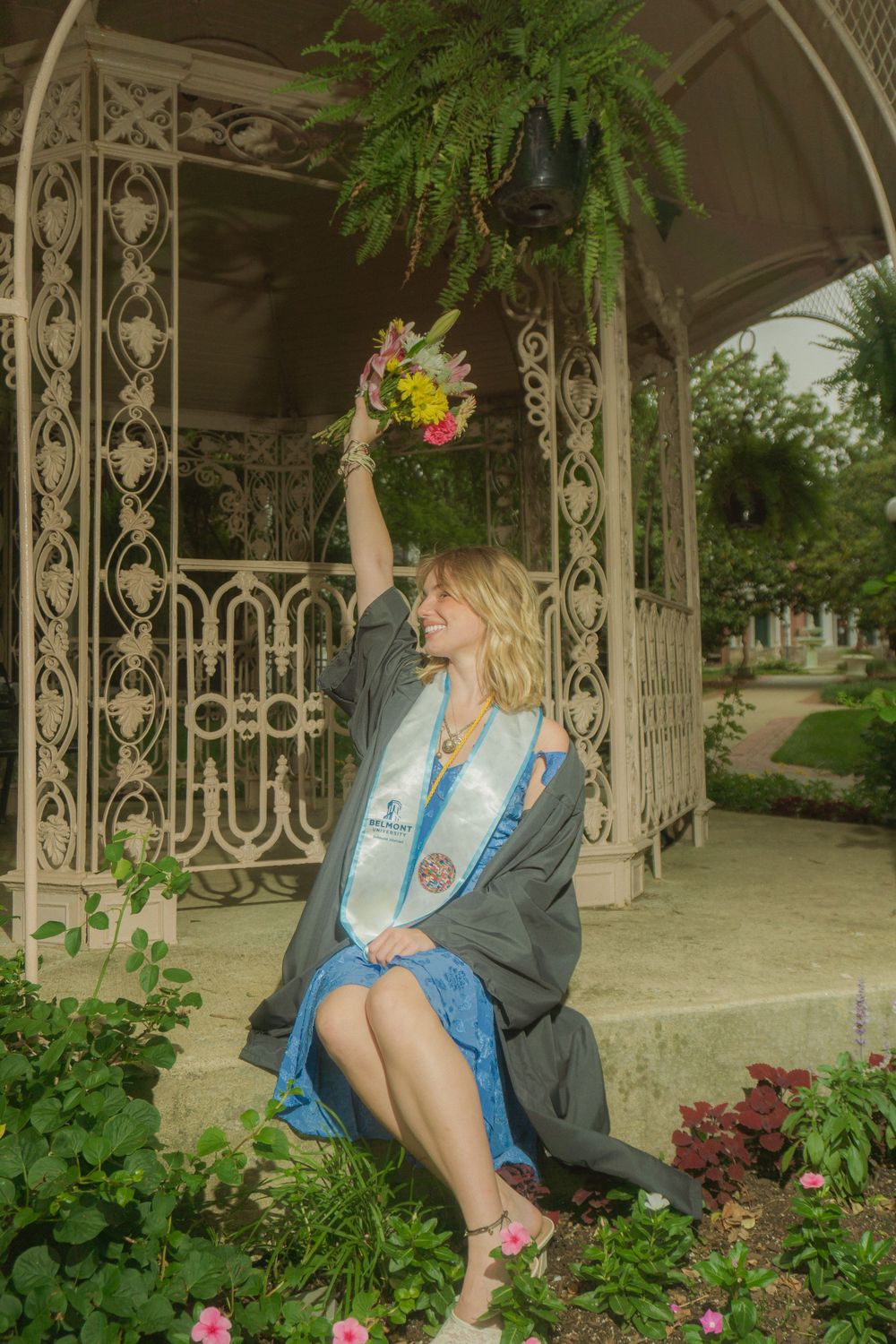 Person in blue dress and dark jacket poses in vintage-style gazebo surrounded by flowers and greenery.