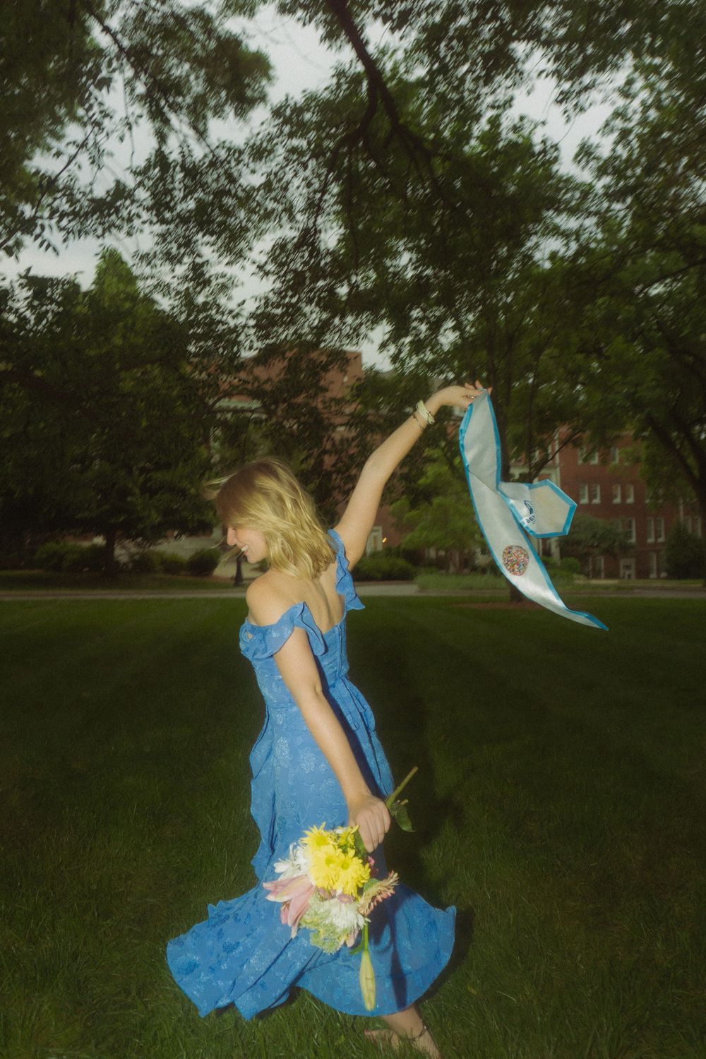 Person in blue dress dances with flowing fabric and flowers in park setting at dusk.