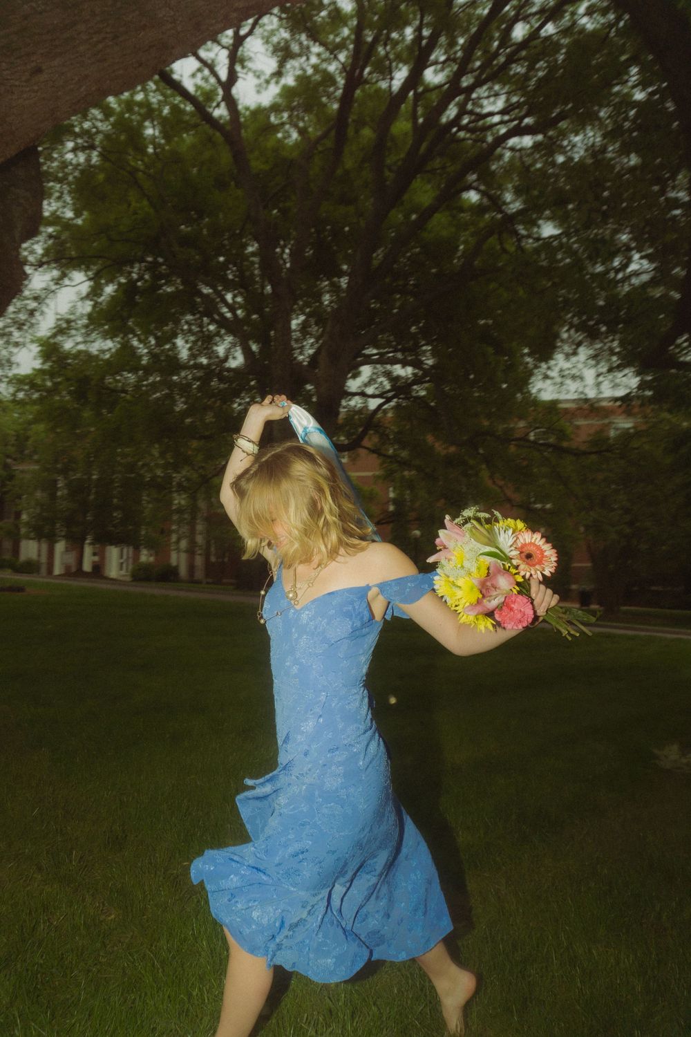 Figure in blue dress spins joyfully with flowers on green lawn with trees in background.