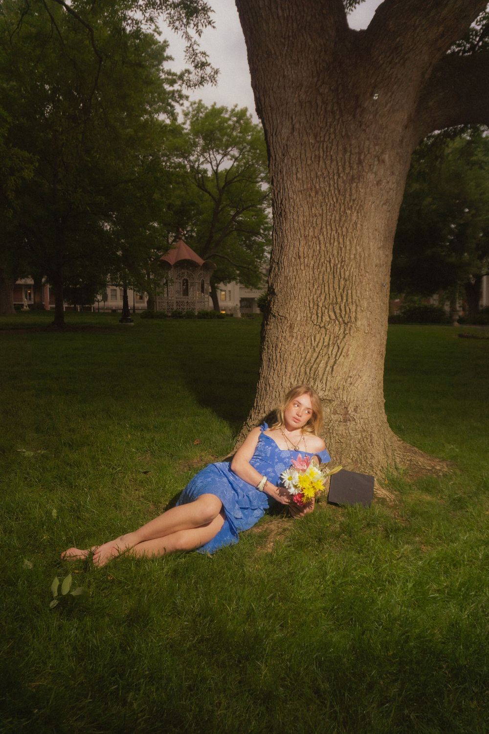 Figure in blue dress reclines against large tree trunk holding colorful flowers.