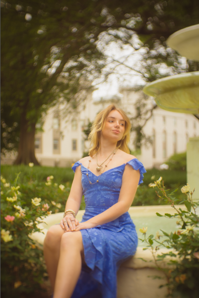 Woman in blue lace dress poses gracefully on garden bench surrounded by blooming flowers and greenery.