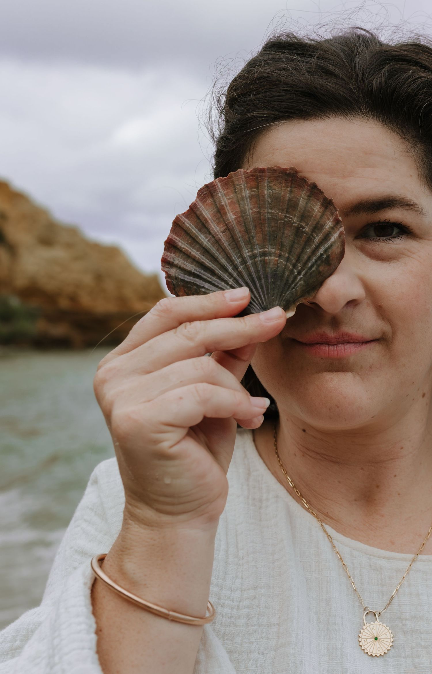 A seashell held up playfully near the ocean with rock formations in the background.