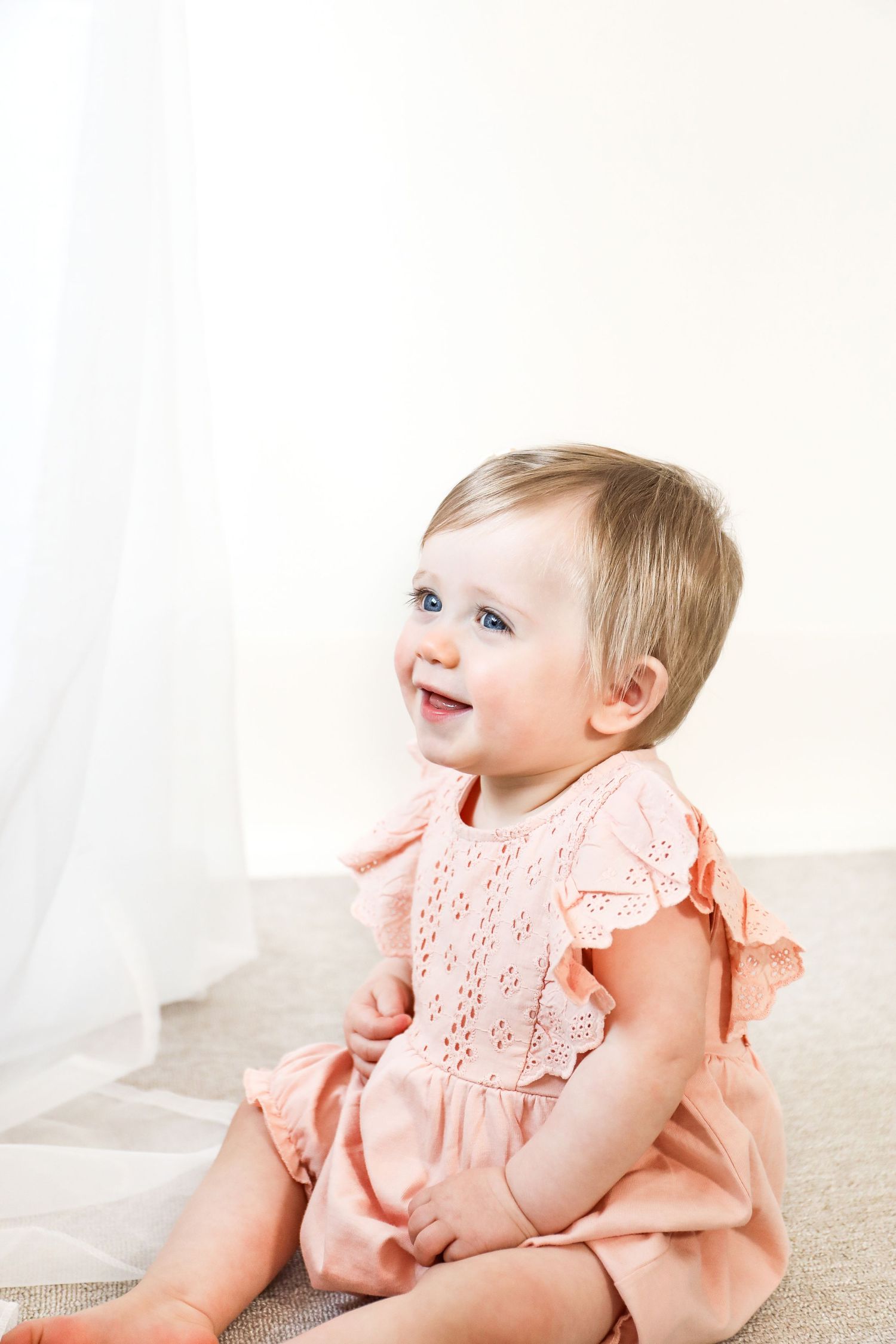 Happy baby wearing a peach-coloured dress sits on floor with a joyful expression, during birthday shoot in Nottingham