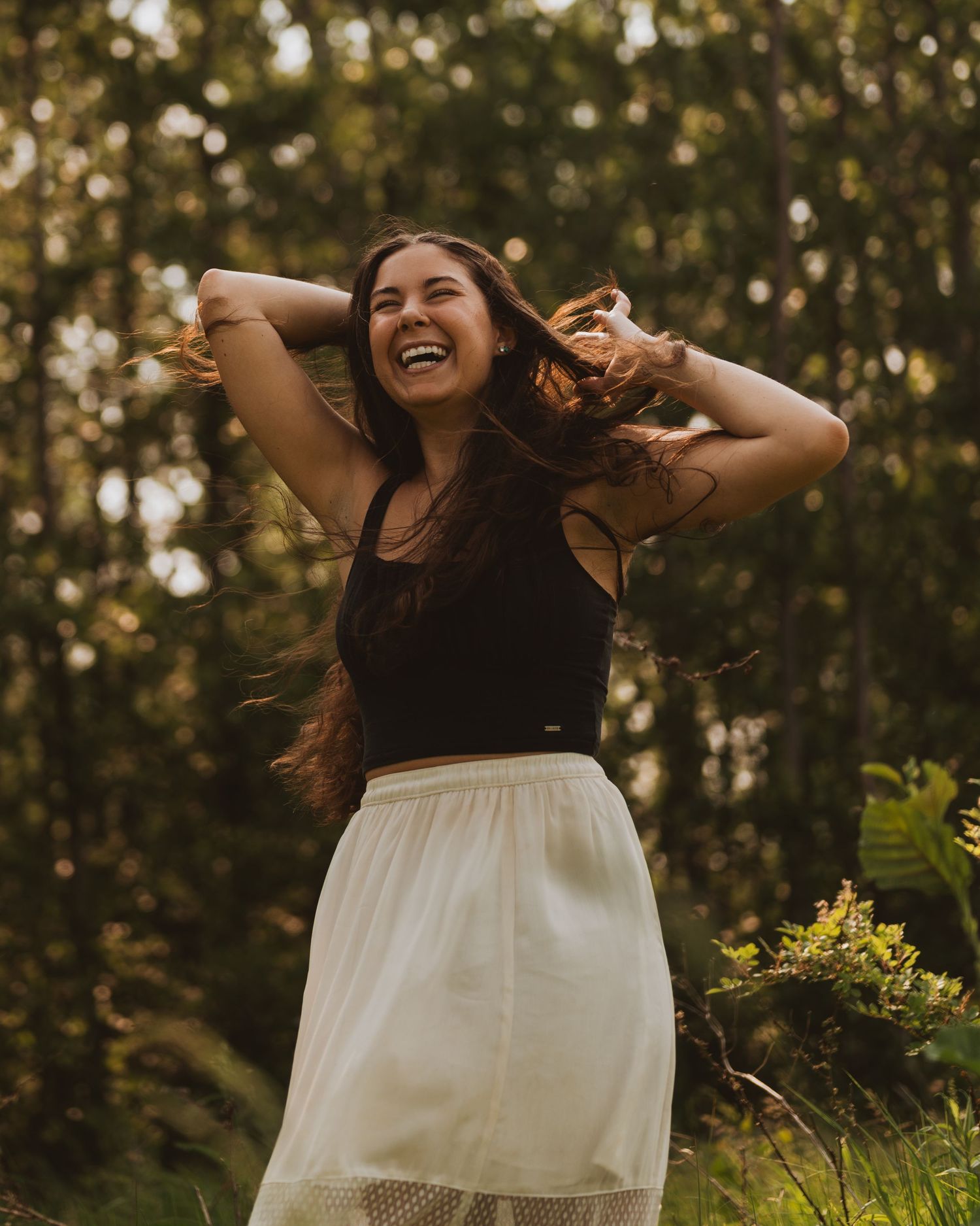 A person wearing a black top and white skirt dances joyfully in a sunlit forest setting during golden hour.