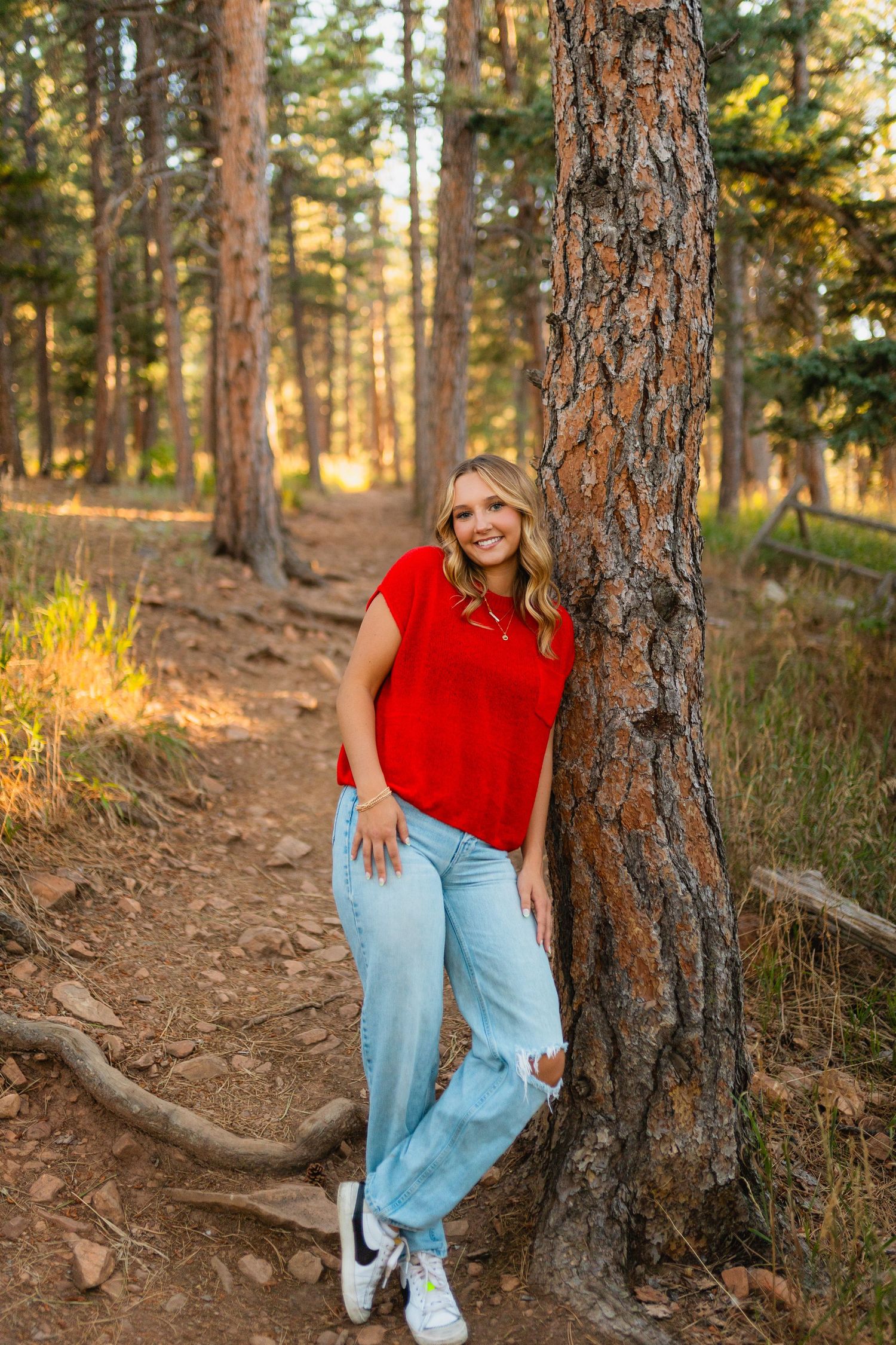 A person in a red sleeveless top and light jeans poses casually against a large pine tree trunk in a sunlit forest.