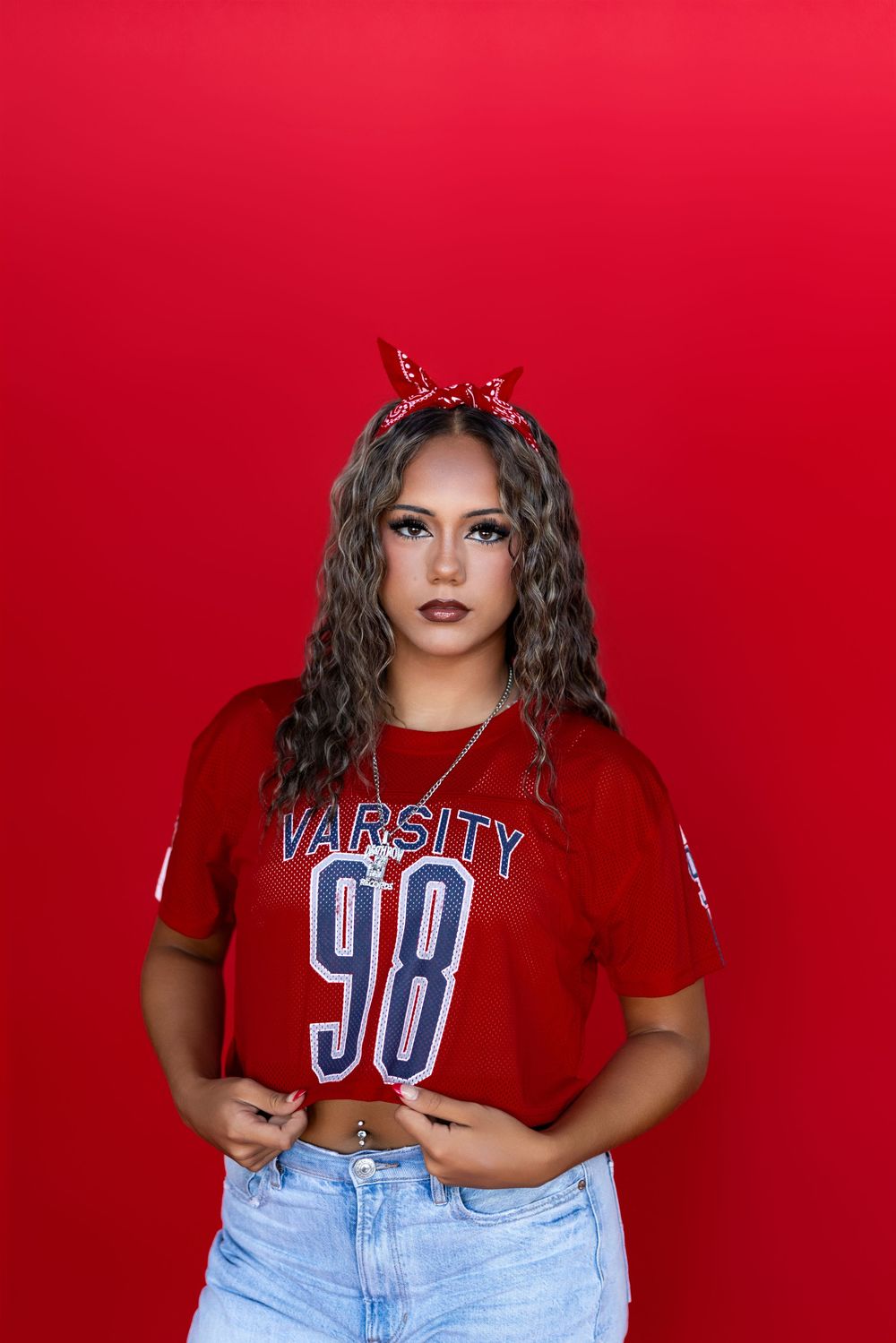 Standing against a red wall wearing a cropped varsity 98 jersey, denim shorts and red glitter horns.