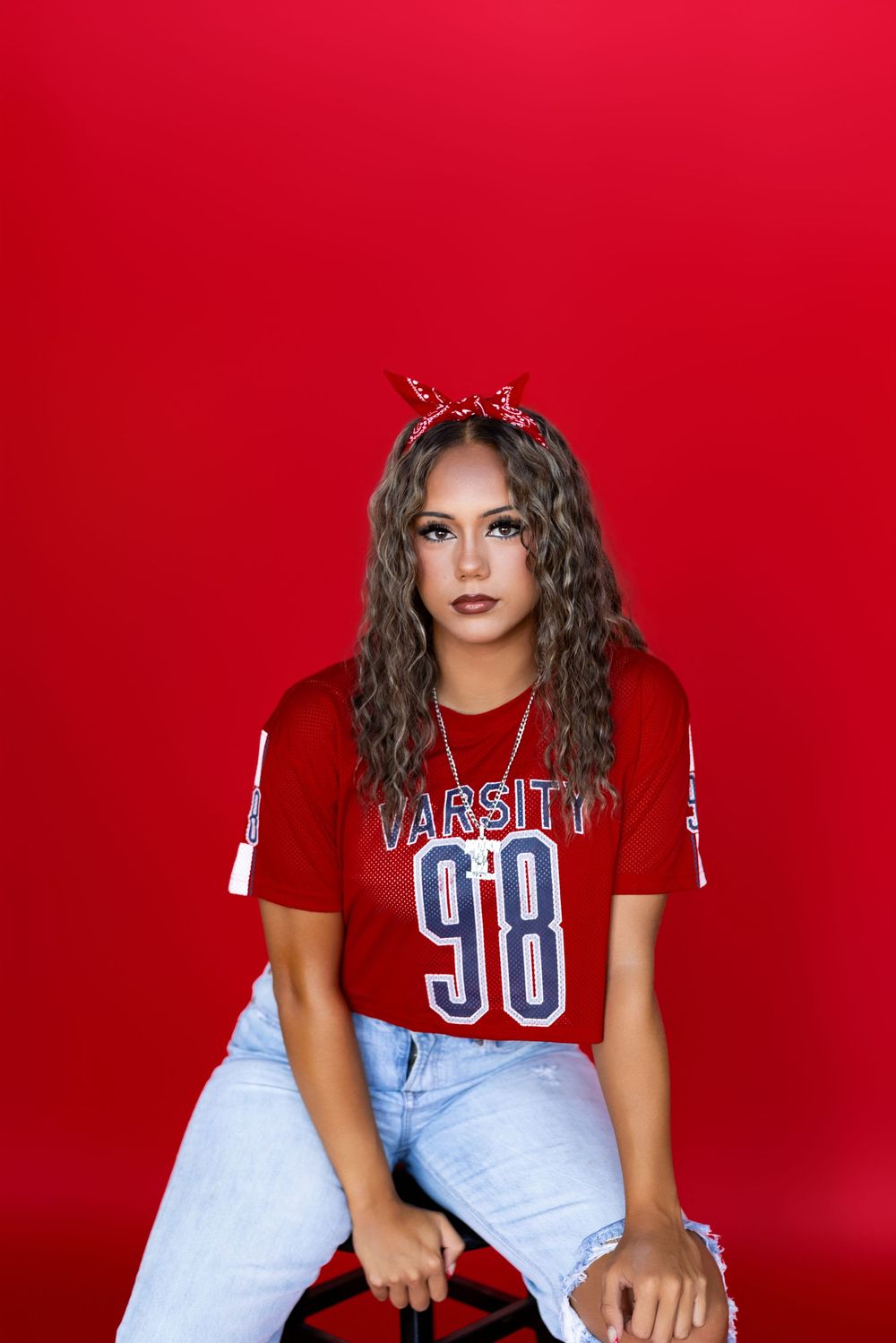 A model in a casual retro varsity jersey numbered 98 poses against a bright red background with a bandana headband.