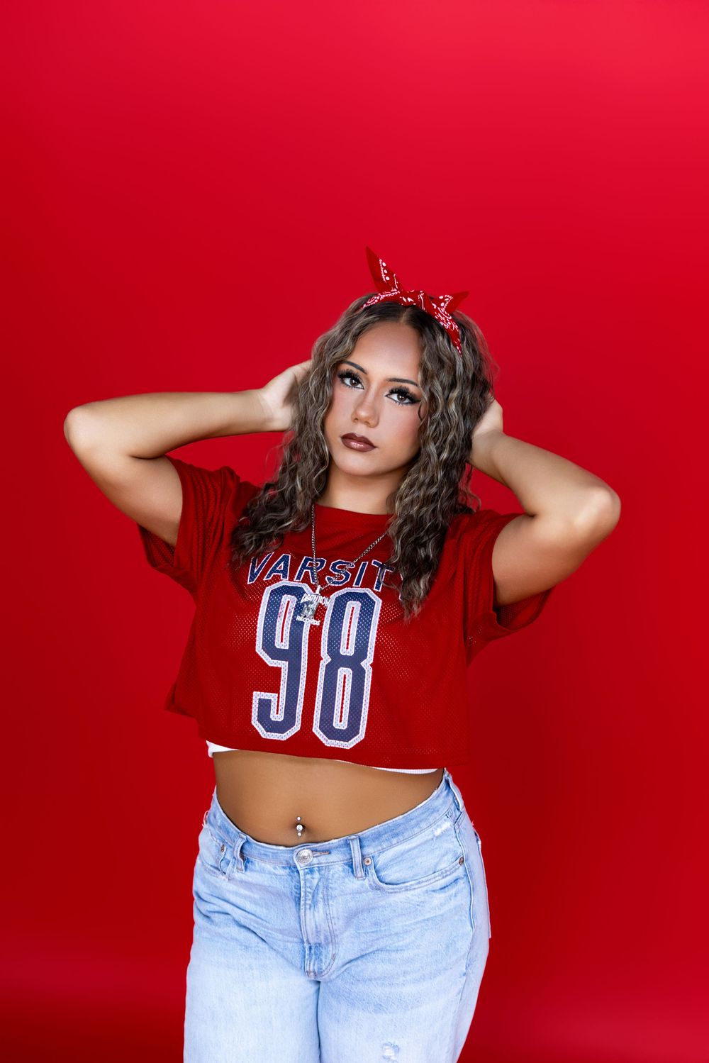 Woman in cropped red sports jersey 98 and light blue jeans poses against vibrant red background.