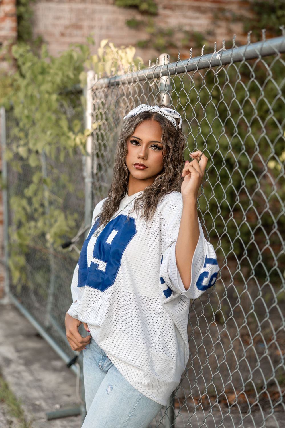 Fashion portrait of someone in a white and blue sports jersey posing against a chain-link fence.