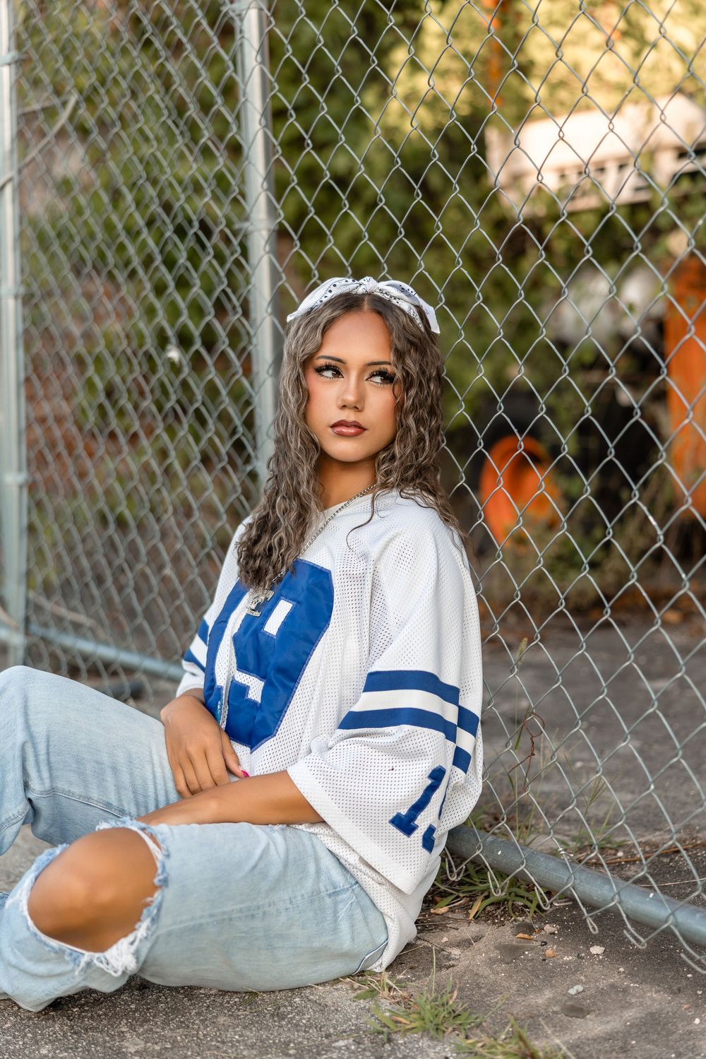 A person wearing a white and blue varsity-style jersey and jeans poses against a chain-link fence at sunset.
