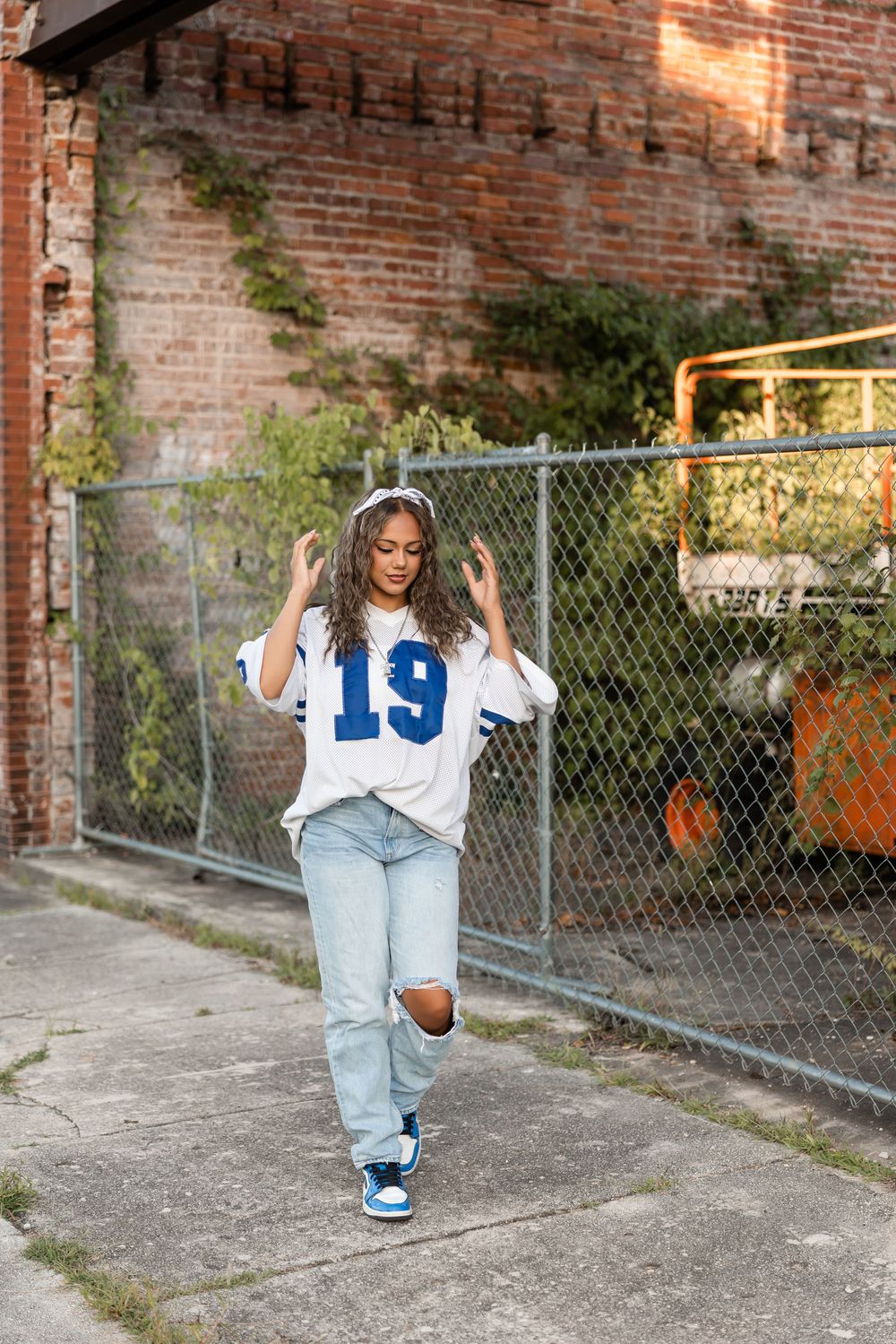 Person wearing a number 19 jersey and ripped jeans poses against brick wall and chain link fence on urban street.