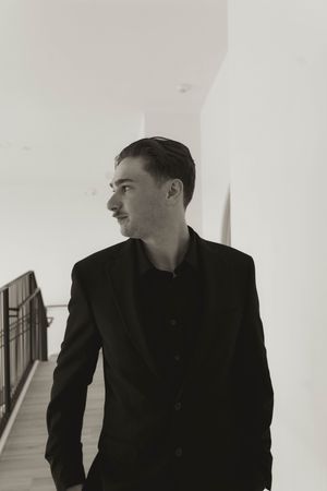 A person in a black suit poses dramatically in a minimalist white hallway.