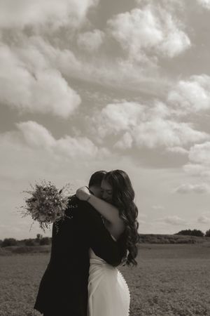 A romantic black and white wedding photo of a couple embracing in a field with dramatic clouds overhead.