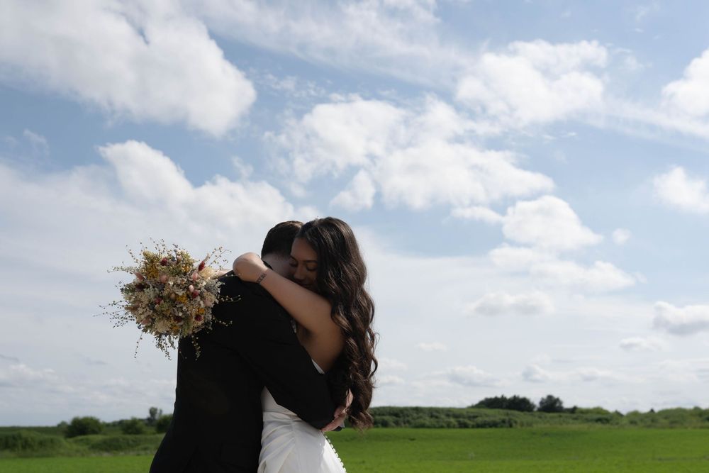 Couple embracing in an open field with dried flower bouquet against cloudy sky.