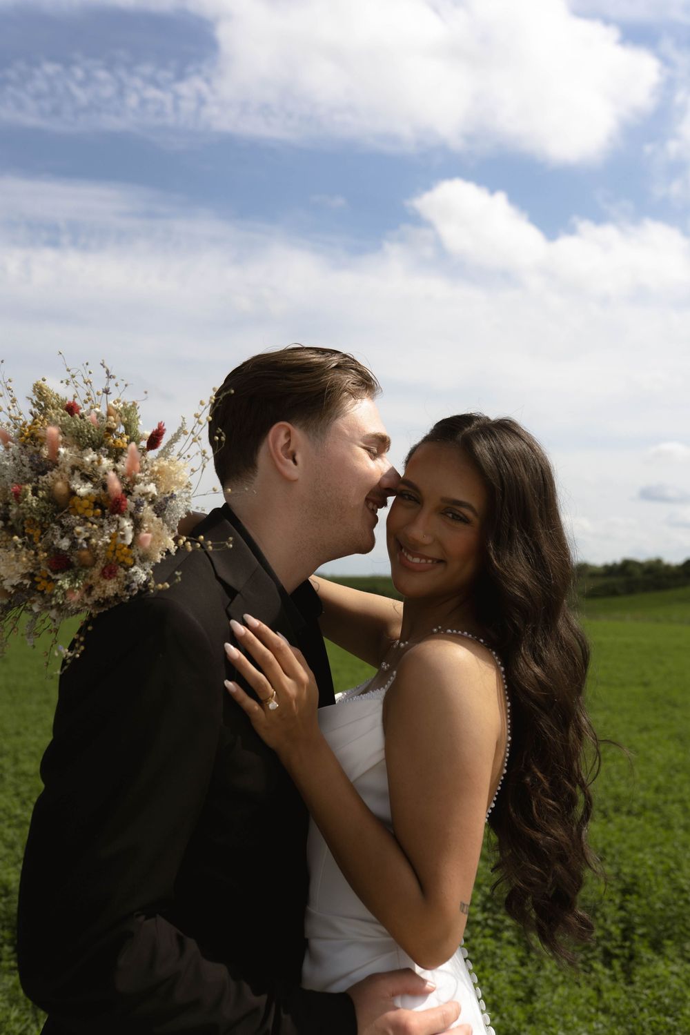 Romantic wedding portrait in a green field with floral bouquet and cloudy sky backdrop.