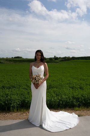 A bride in a flowing white dress poses with her bouquet against a lush green countryside backdrop.