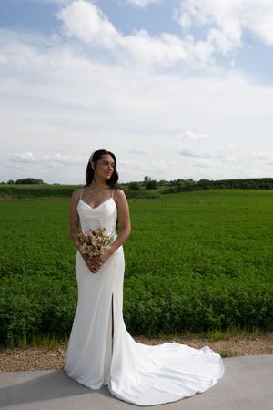 A bride stands elegantly in her white gown with a trailing train on a path beside green fields.