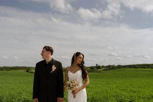 A couple in wedding attire stands together in a lush green field under cloudy skies.