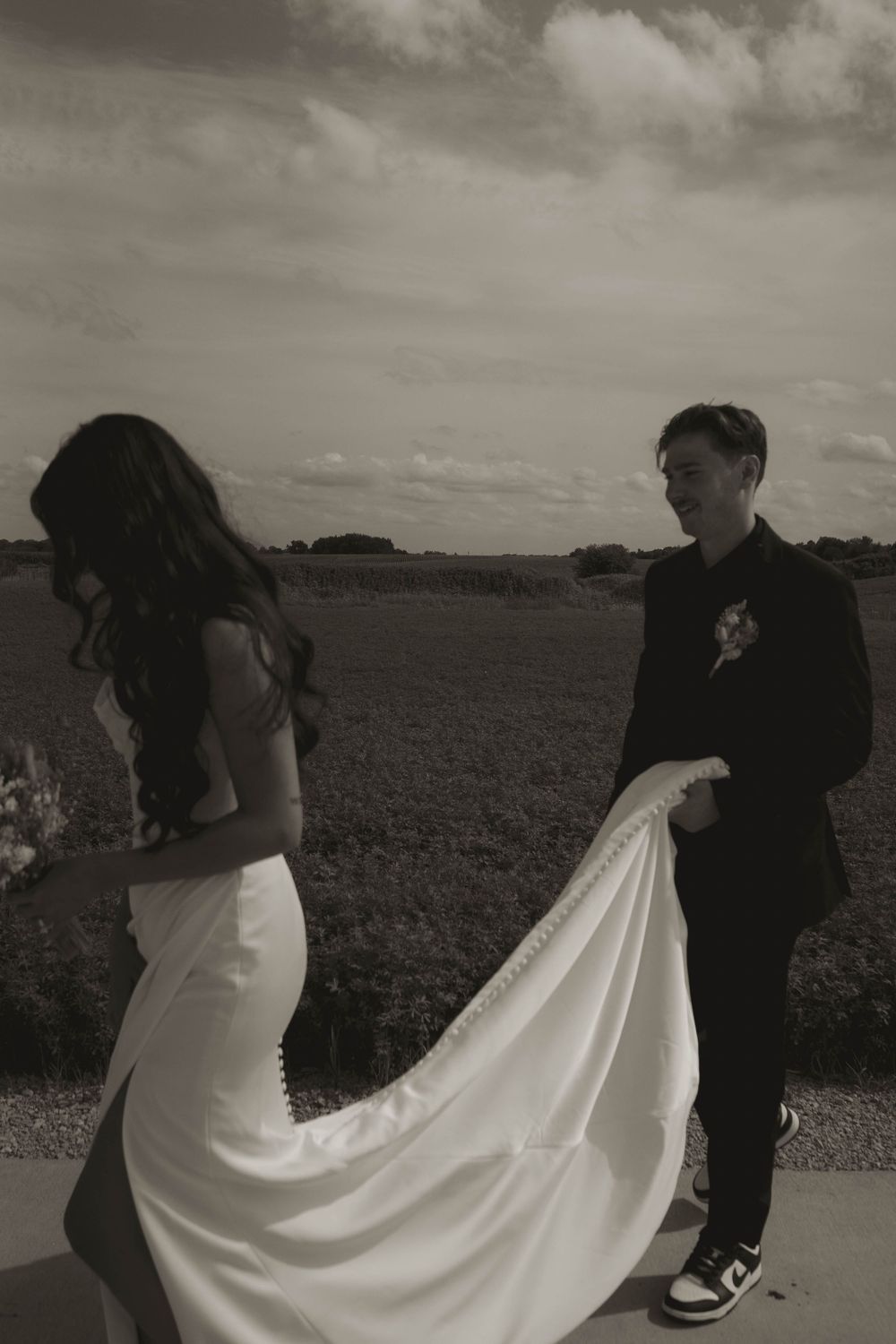 Black and white wedding photo with flowing dress train in rural landscape.