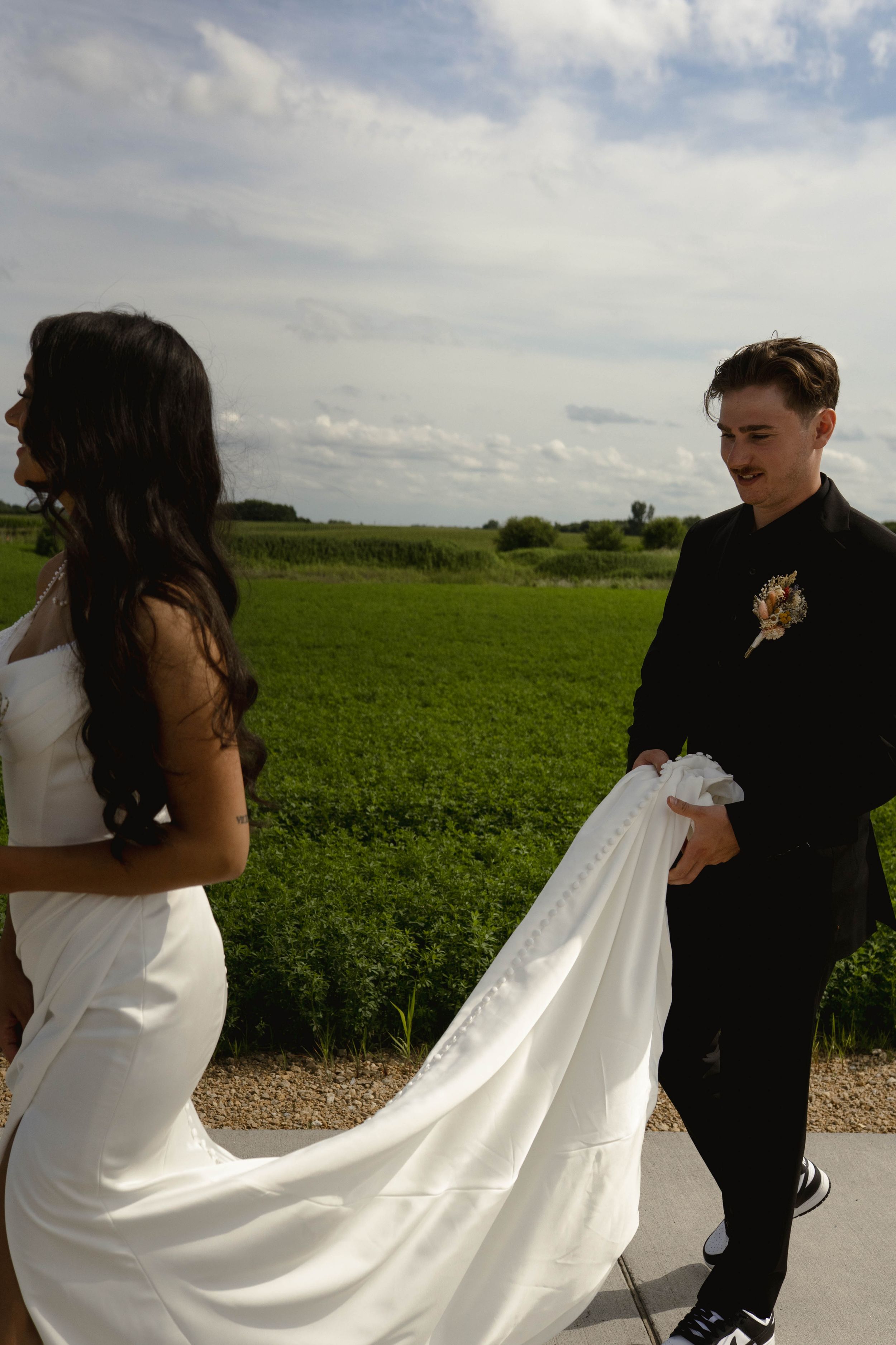 Wedding couple on path with flowing white dress train against green countryside.