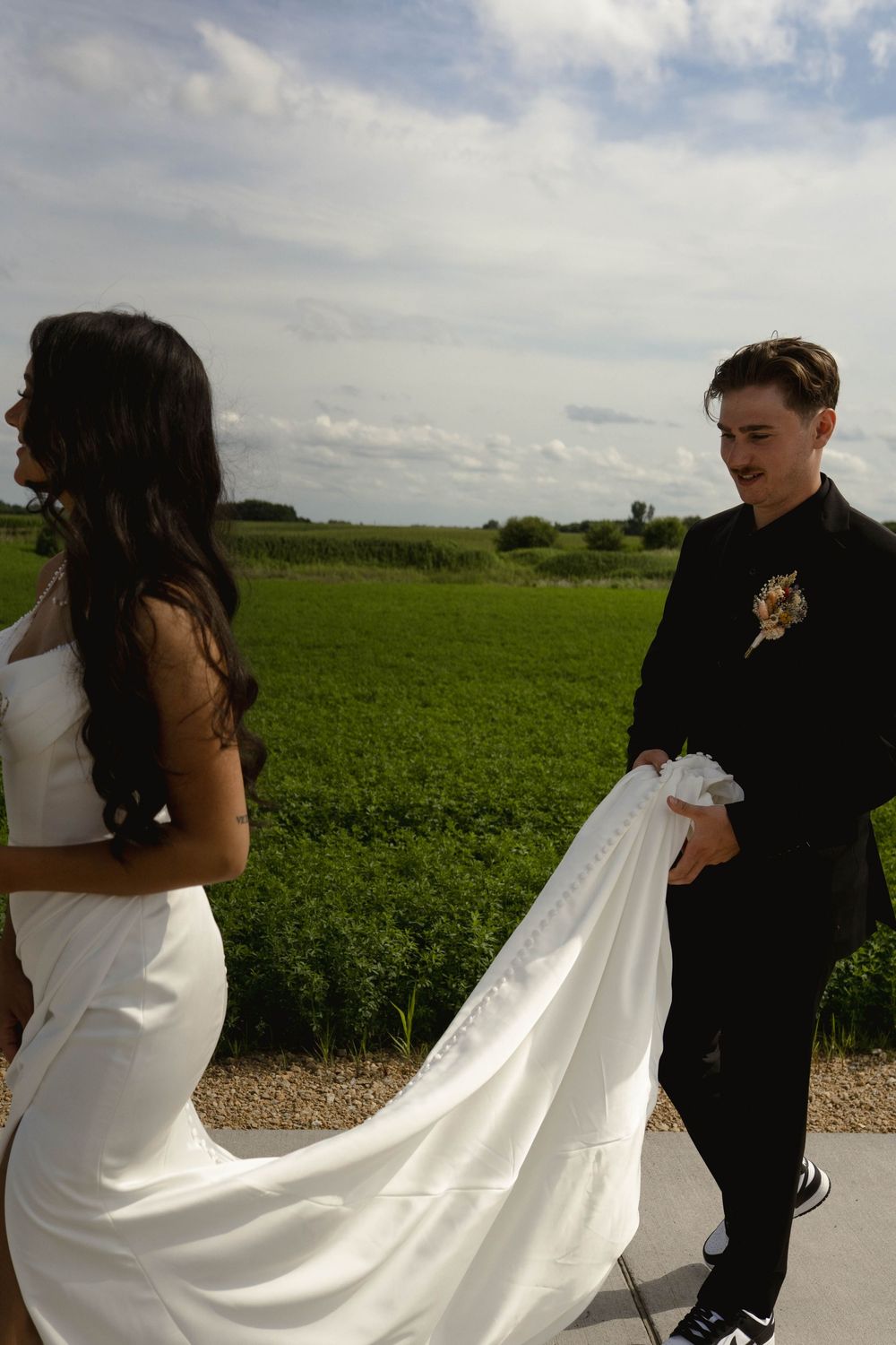 Wedding couple on path with flowing white dress train against green countryside.