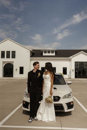 A couple in wedding attire poses in front of a white building with their car on a sunny day.