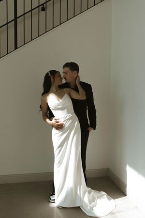 A couple in wedding attire shares an intimate moment in a minimalist white hallway with stairs.